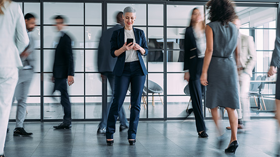 A woman wearing professional clothes is standing among several walking professionals while looking at her mobile device.