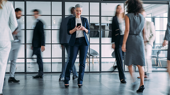 A woman wearing professional clothes is standing among several walking professionals while looking at her mobile device.