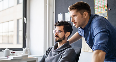 Two men, one seated and the other standing, are looking at a computer monitor.