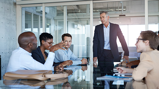 A man wearing professional clothes is standing at the head of a meeting table talking with several adults.