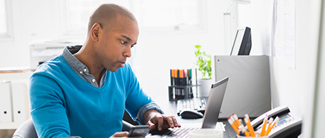 A man wearing professional clothes is typing at a computer and holding a mobile device while seated at a desk. 