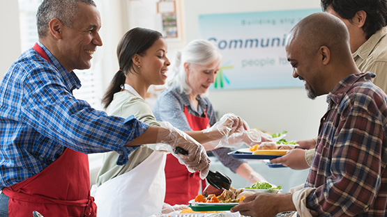 Three adults standing in a row are wearing aprons and serving food to two other people.