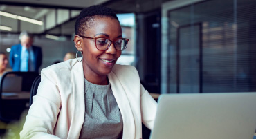 A woman wearing professional clothes is smiling while sitting down working on a laptop.