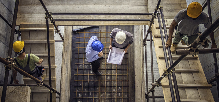 Two workers on a job site wearing helmets look at blueprints while two other workers in helmets walk across nearby rafters.
