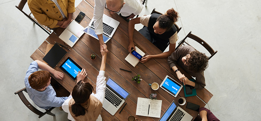 A group of men and women are seated at a table with laptops and tablets-one man and woman shake hands across the table while the rest look on.