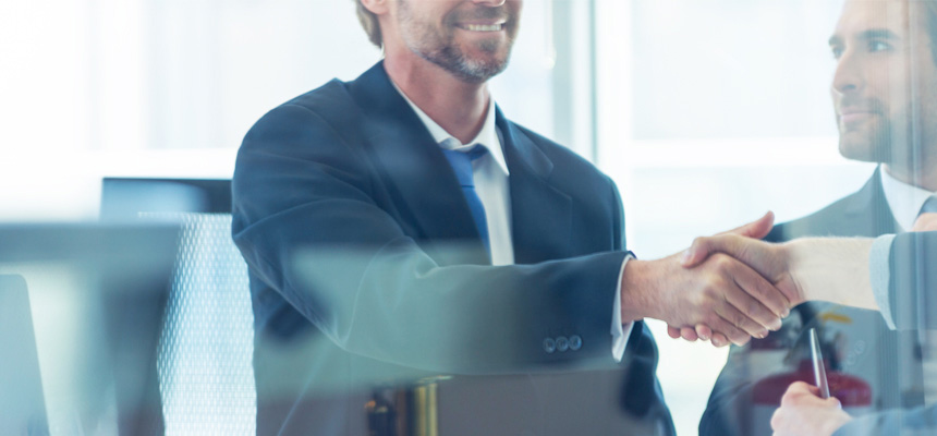 Two seated people wearing professional clothes are shaking hands while a third professional male looks on.