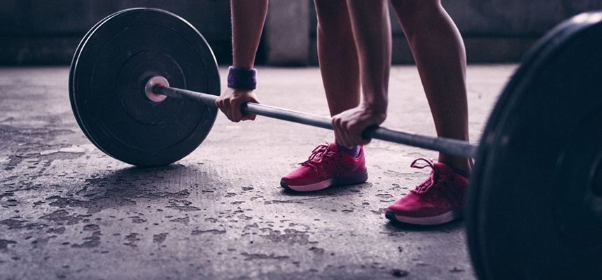A person wearing gym shoes has hands positioned to lift a barbell that is placed on the floor.