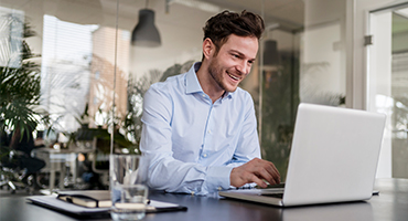 A man wearing professional clothes is sitting at a table working on a laptop.