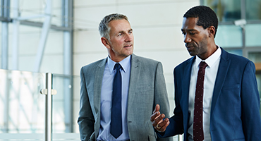 Two men wearing professional clothes walking down a hallway talking with each other.