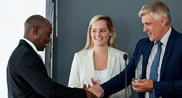 A man wearing professional clothes stands at a podium shaking hands wtih another man in professional clothes while a professionally dressed woman stands nearby.