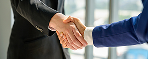 Two people wearing professional clothes shaking hands in front of a tall window pane. 