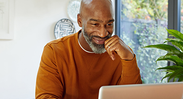 A smiling middle-aged professional looks at a laptop with one hand to their chin, representing the ease and confidence clients experience with HM Insurance Group's efficient claims processing and online tools.