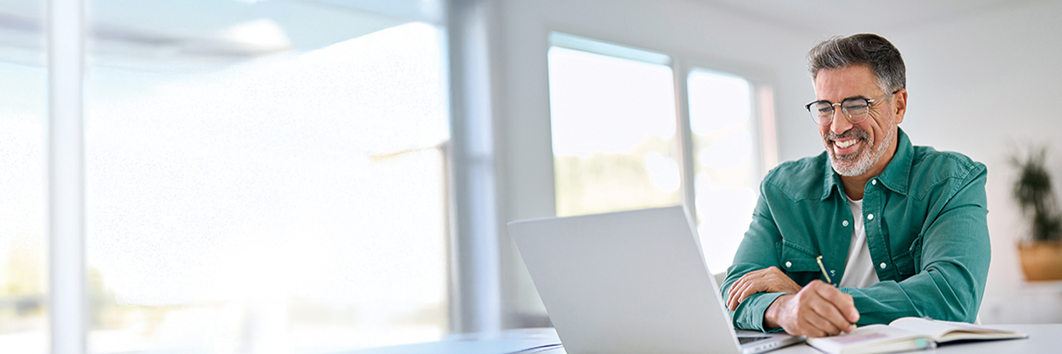 A middle-aged professional smiles while working at a laptop and taking notes, embodying the focused and supportive partnership HM Insurance Group extends to its clients in financial risk protection.