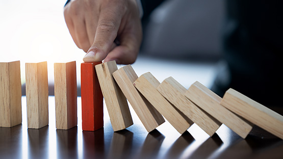 A person's hand stops a red domino from toppling other dominos, symbolizing how HM Insurance Group prevents financial risk for self-funded employers.