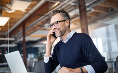 A man is sitting at a computer smiling while talking on the phone