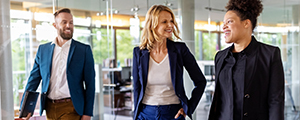 A female insurance broker wearing professional clothes walks down a hallway while talking with another female..