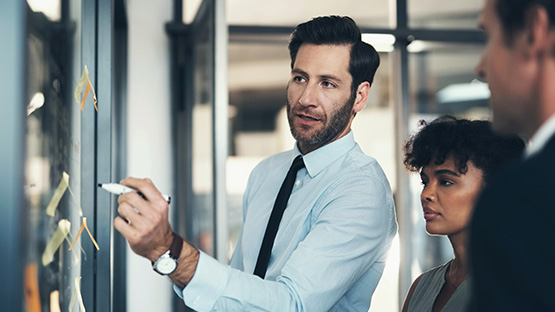 A standing male insurance broker wearing professional clothes points to a whiteboard while a standing man and woman follow along.