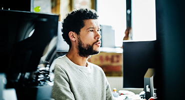 A seated man, surrounded by several work stations, looks at his computer monitor.