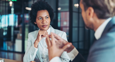 A male insurance broker wearing professional clothes and female medical professional are talking while seated near an indoor plant.