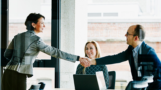 A female insurance broker wearing professional clothes and man stand to shake hands while a seated woman smiles at them. 